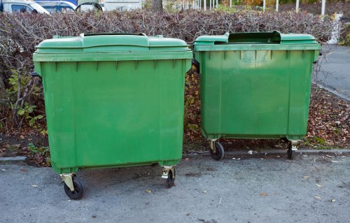Two large green commercial rubbish bins with smooth plastic surfaces and slightly curved lids, positioned next to each other on a paved surface outdoors. The bins have four small black wheels at the base, with the left bin slightly larger and taller than the right. They are situated against a background of bushes with dark green and brown foliage, indicating an autumn or winter setting, on a paved area possibly part of a commercial or residential vicinity in Redbridge, London. The lighting appears natural and overcast, casting minimal shadows. These bins are typical of waste management services provided by Office Clearance Redbridge for local rubbish collection and disposal needs, with a focus on general waste or rubbish removal from nearby office or residential premises.