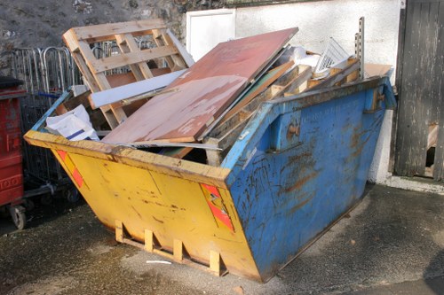 A large, rectangular metal skip with a yellow front panel and blue sides, positioned outdoors on a paved surface adjacent to a concrete wall and black wooden gate in Redbridge. The skip is filled with various construction debris, including wooden pallets, metal panels with a reddish-brown finish, and miscellaneous lightweight materials. Some of the wood appears weathered, with visible grain and rough edges, while the metal objects include perforated sheets and upright pieces. The skip's open top reveals the cluttered contents, and the surrounding environment suggests a temporary waste collection area for rubbish removal services in the local area. The ground beneath the skip is damp, indicating recent cleaning or rain, and the weather appears overcast, providing diffuse lighting across the scene. This visual context aligns with professional waste management operations in London suburbs like Redbridge, where rubbish collection and site clearance are common activities carried out by companies such as Office Clearance Redbridge.