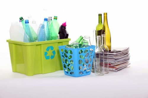 In the foreground are two plastic containers used for recycling or waste collection, with a large lime green bin positioned to the left holding several transparent plastic bottles in shades of blue, green, and clear glass. To the right, a smaller light blue basket with circular cut-outs contains more glass bottles, some green and others clear. Behind these containers, there are additional glass bottles, including a tall yellow bottle and a shorter green one, placed on a surface along with a neatly stacked pile of newspapers or magazines. The background appears to be a plain, bright white setting, emphasizing the waste items and containers, which are all arranged in an organized manner suitable for professional rubbish removal services. The scene suggests a focus on separating and managing recyclable waste items, consistent with the offerings of Office Clearance Redbridge, serving the local postcode area, with a subtle emphasis on environmentally responsible rubbish disposal.