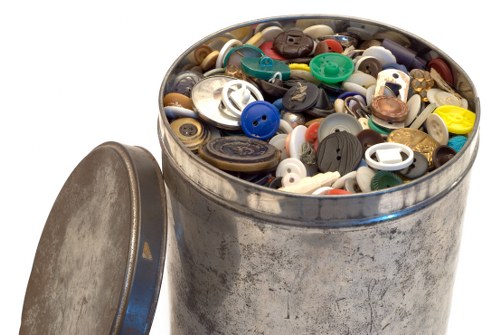 A close-up image of a round metal container filled with a large assortment of assorted buttons in various sizes, shapes, and colours, including black, white, blue, green, brown, and yellow. The container is silver with a slightly weathered appearance, and its lid, which is leaning against the side, appears to be made of the same metal with a dull finish. The buttons are densely packed, some overlapping each other, and are situated on a plain, white background. The image is related to rubbish removal services such as those offered by Office Clearance Redbridge, focusing on the disposal or collection of small household items like buttons, which could be part of a larger clearance or decluttering project in the Redbridge area, postcode suggested by the context. The lighting is even, highlighting the textures and colours of the buttons clearly, with a focus on neatness and simplicity vital for accessibility and visual clarity. The overall tone is neutral and professional, fitting for a service involved in waste management or rubbish clearance, emphasizing the variety and compact arrangement of small recyclable or disposable items.