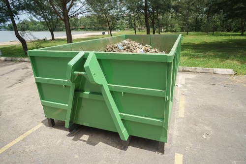 A large, industrial green skip bin filled with mixed waste materials, including paper, plastic, and organic debris, situated on a paved parking area with yellow lines. The bin is positioned near a grassy area with trees in the background, and a pathway or river visible beyond the trees, indicating a park or outdoor space. The skip has a robust metal construction with horizontal reinforcement panels and an angled lever mechanism on one side for opening the lid. The environment appears to be outdoors during daylight, with natural light illuminating the scene, suitable for rubbish removal services in Redbridge or nearby areas. This image exemplifies the type of waste collection equipment used for clearing rubbish from commercial or residential sites, as managed by Office Clearance Redbridge.
