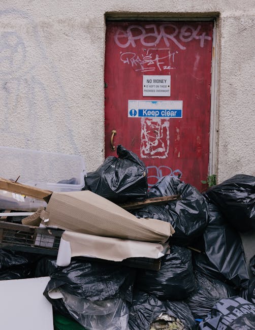 A collection of overflowing rubbish bins and scattered waste on a paved area in an urban environment. The scene includes a large grey mixed paper and card bin with its lid open, overflowing with vario