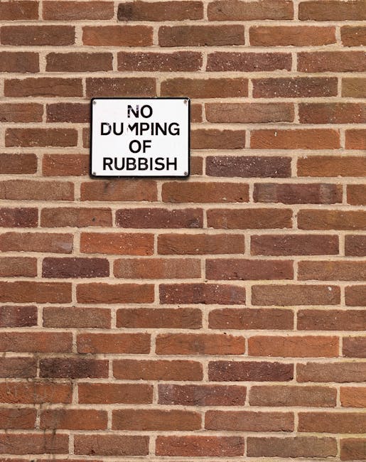 A close-up view of a rectangular white metal sign mounted on a red brick wall, which reads 'NO DUMPING OF RUBBISH' in bold black letters. The sign shows slight wear, with some areas of minor rust or d