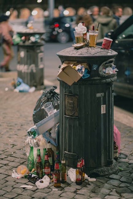 A green outdoor waste bin with a rounded lid, positioned on a metal pole in a grassy area with a dirt pathway in the background. The lid is partially open, revealing assorted rubbish including paper a